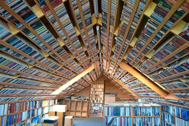 Photo of the a different angle of the top floor of a house with the slated roof and walls all but covered in books in bookcases. 