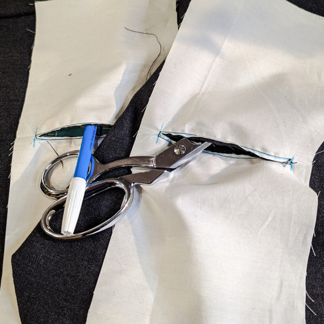 A fabric pen and sewing scissors sticking out of slashed holes in white pocket fabric. The holes, surrounded by black stitching, go through the pocket fabric and the wool fashion fabric underneath.