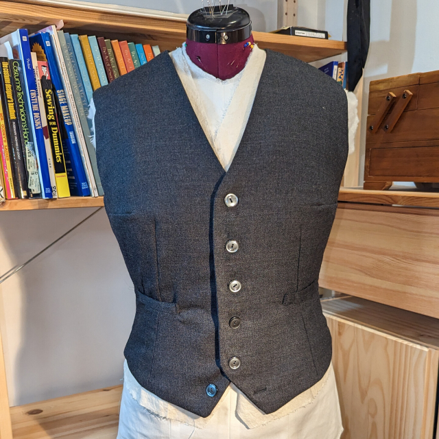 A custom tailored grey wool waistcoat with pearlescent grey buttons, on a custom padded dress form in front of a bookcase