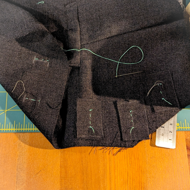 Center front of a heathered charcoal waistcoat piece, with rectangles of the same fabric tacked in place over the button hole marks with bright teal thread