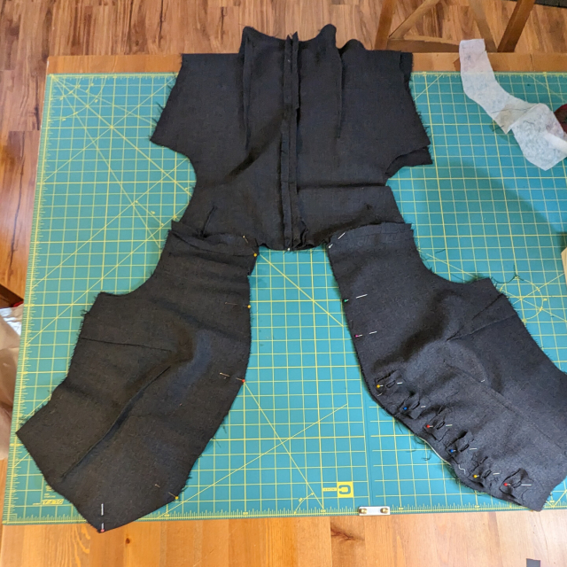 A dark grey waistcoat in progress, spread out on a sewing table. The lining, face up, is pinned to the outer fashion fabric at the neck, lapel, and center front