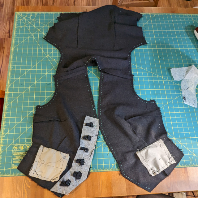 A waistcoat in progress sitting on a workbench. Teal basting lines show against the grey fabric.