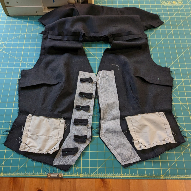 A grey waistcoat in progress, inside-out on a green work table. Black stitch lines are visible along the white interfacing at the front.