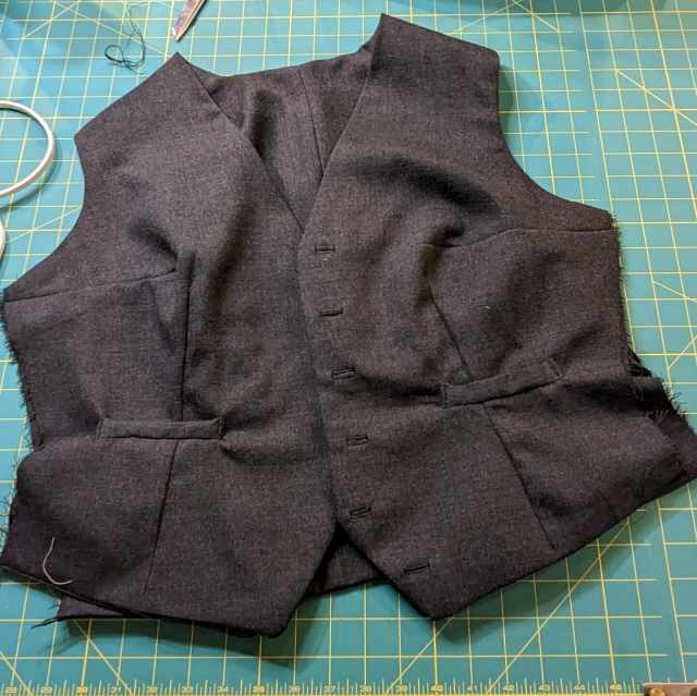A grey waistcoat in progress on a green work mat. The side seams are still open.