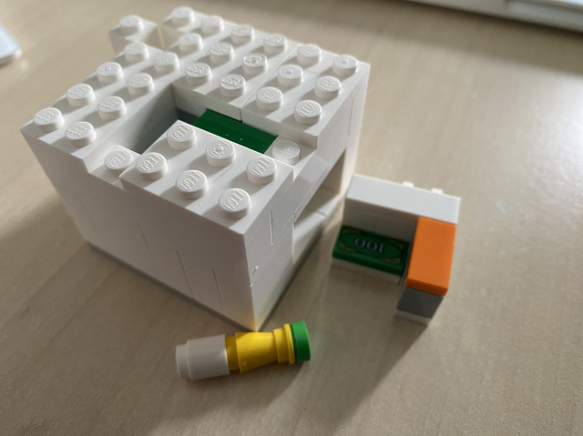 The same cube of Lego except now a drawer has been removed to reveal a $100 tile and a staff of green, yellow and white bricks sits beside it