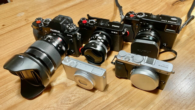 a collection of fujifilm cameras on a table, some with large bulky lenses, others with small lenses or built in retracted lenses.