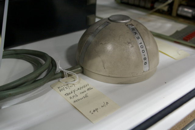 A semisphere on a desk top, about the size of half a tennis ball, with the flat size down. It’s beige and scruffy. A green cable is attached at the based and curled up on the left. On the top of the sphere is a silver button. A label on the sphere reads "RKS 100-86"