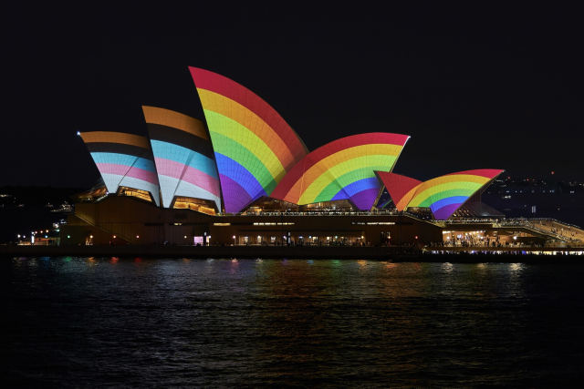 A photo of the Sydney Opera House taken from the Overseas Passenger Terminal. Projected on its sails are the colours of the Progress Pride Flag.
