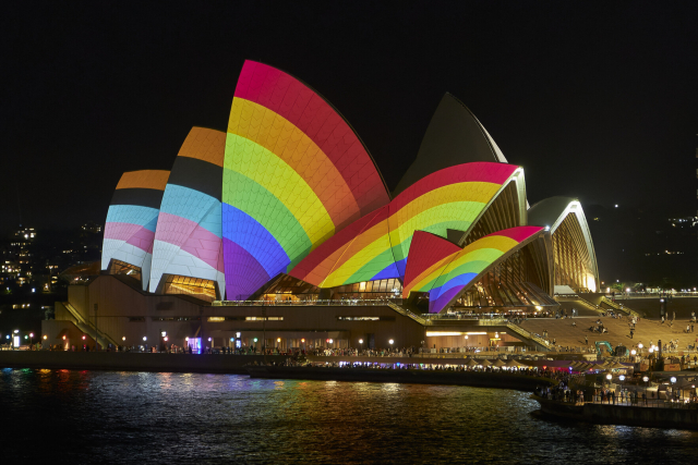 A photo of the Sydney Opera House taken from the Cahill Expressway. Projected on its sails are the colours of the Progress Pride Flag.
