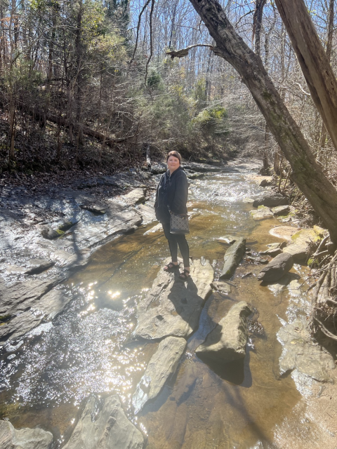 Woman standing on flat rock in middle of stream with trees on the sides, sunny ￼