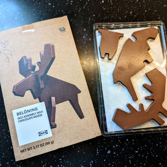 A flat cardboard box with an image of a very stylized moose made from three flat pieces of chocolate — a giant set of antlers, a profile of a head and body with characteristic snout, and a third piece comprising the front legs, meant to mount perpendicular to the body so it can stand. The box has a large label that reads “Belöning, self-assembly milk chocolate moose” with the IKEA logo. Next to it is the tray pulled from the box holding the pieces of chocolate, definitely not assembling itself while I watch.