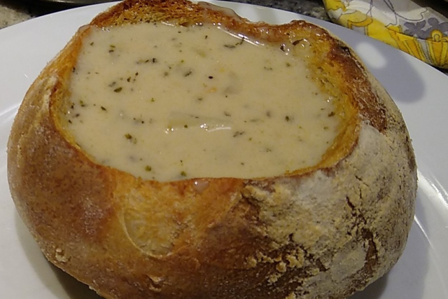 A hollowed out round loaf of bread is filled with creamy, herbed chowder and is on a plate