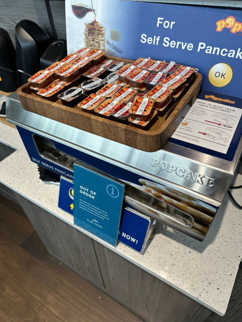 The self-serve pancake machine has a tray of jam pots on top of it, and a sign in front of it saying “out of order”