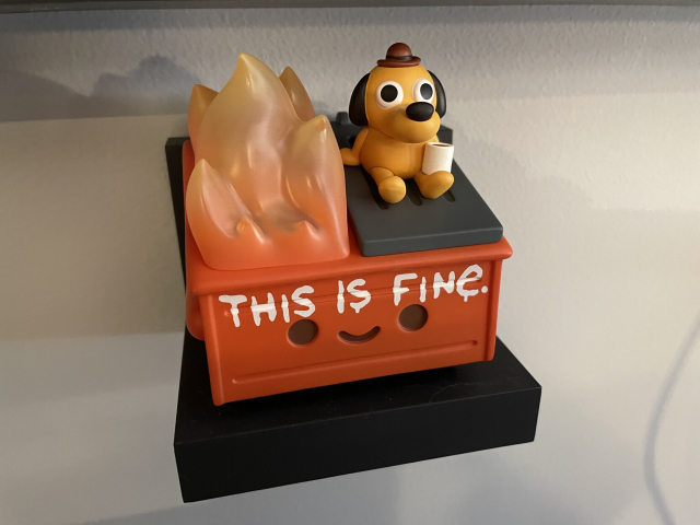 A vinyl figurine of an anthropomorphic, smiling, burning garbage dumpster with the “this is fine” dog sitting atop it. 