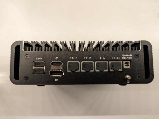 Back panel of a small fanless computer with four Ethernet ports and two USB-A ports with black silicone dust plugs in them.