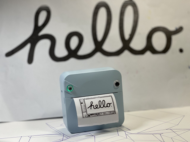 A small handheld thermal wireless printer in light blue with paper printed that says hello in cursive on it coming out. In the background is the cursive word hello. 