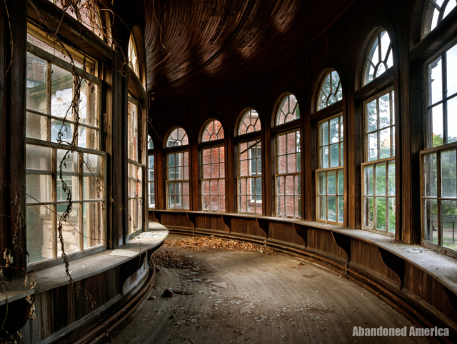 A beautiful; curved hallway lined with arched windows, at the bottom of which is a rim large enough to sit on. Vines have started to crawl through the windows and leaves are scattered on the floor