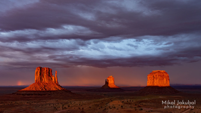 A dramatic scene looking out over a broad desert plain with three massive, bright orange stone towers. The sky is dark and stormy and the foreground is in shadow. There is a small glimpse of rainbow to the left of one of the towers. The towers themselves are bathed in deep red-orange sunset glow. 