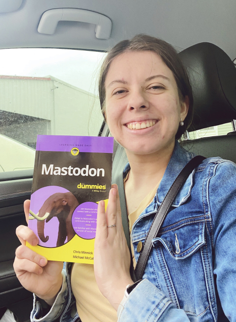 A photo of a white woman sitting in a car, wearing a t-shirt and a jean jacket. She is holding a book titled “Mastodon for dummies” by Chris Minnick and Michael McCallister 