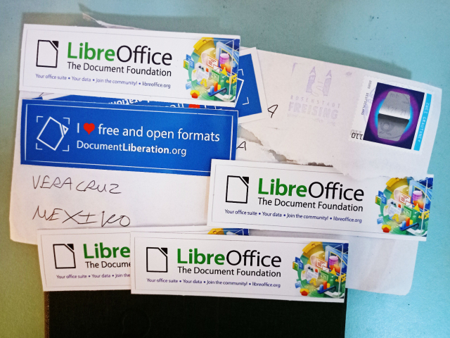 Foto: encima de un sobre postal abierto, varias pegatinas rectangulares en dos diseños.
1) La imagen "splash" que aparece al iniciar LibreOffice 7 (fondo blanco, el imagotipo de la marca y una ilustración  estilo "clipart", que alude a las funciones de oficina, en el borde derecho).
2) En fondo azul rey, con trazos blancos, el icono de LibreOficce "en la mira", sobre el lado derecho del rectángulo, en el resto, se lee "I ❤️ free and open formats" y, debajo de esa frase, "DocumentLiberation.org".
