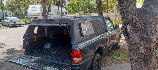 View from the rear passenger side of the Ranger. A poorly painted black camper shell sits on the bed. The shell hatch is open and the tailgate is down. All the visible windows on the shell are painted over, mostly flat black.