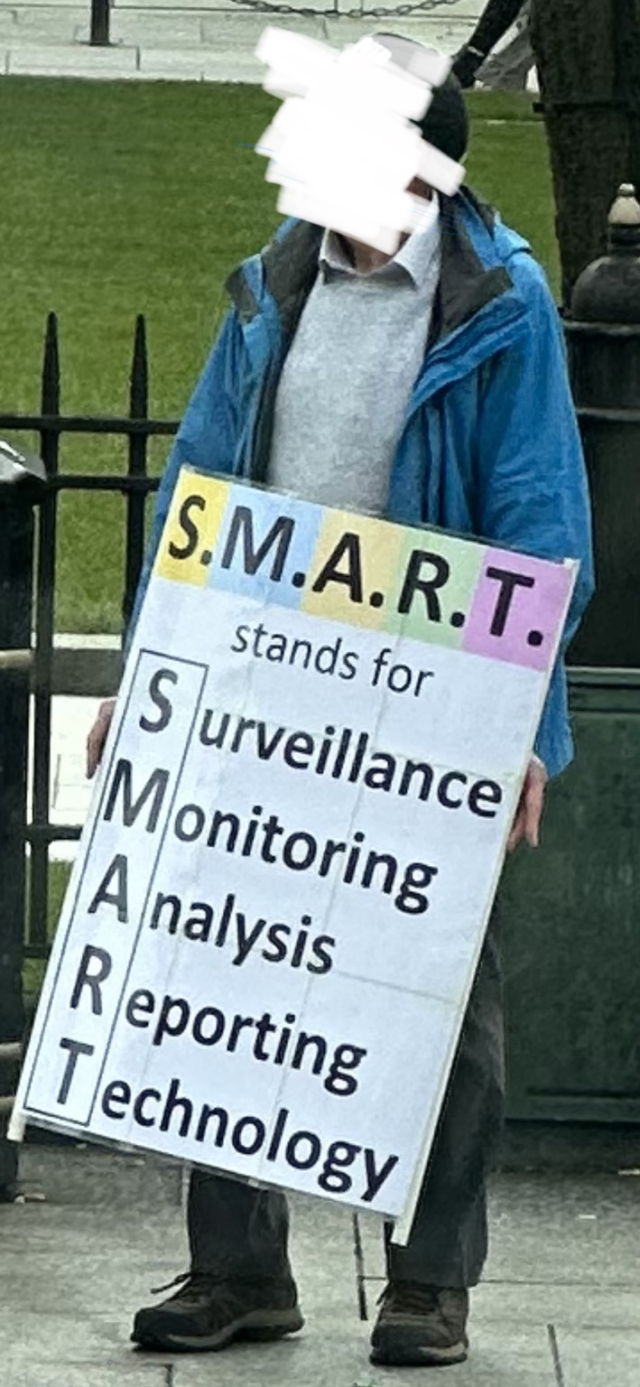 a protestor holding a handmade sign. the sign says “s.m.a.r.t. stands for surveillance, monitoring, analysis, reporting technology”. 