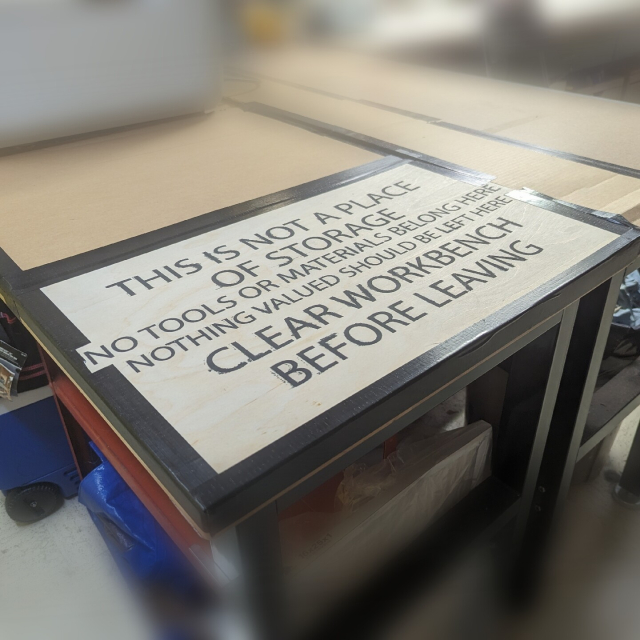 A stencil painted plywood sign on a cardboard covered workbench reads "This Is Not A Place Of Storage / no tools or materials belong here / nothing valued should be left here / CLEAR WORKBENCH BEFORE LEAVING"