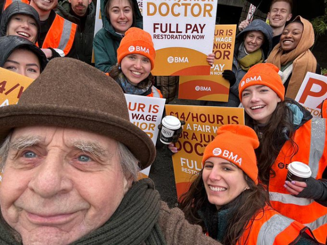 Photo of a picket line with many picketers wearing orange BMA hats and holding signs demanding full pay for junior doctors. Sylvester McCoy, in a brown trilby, stands in front, taking a selfie. 