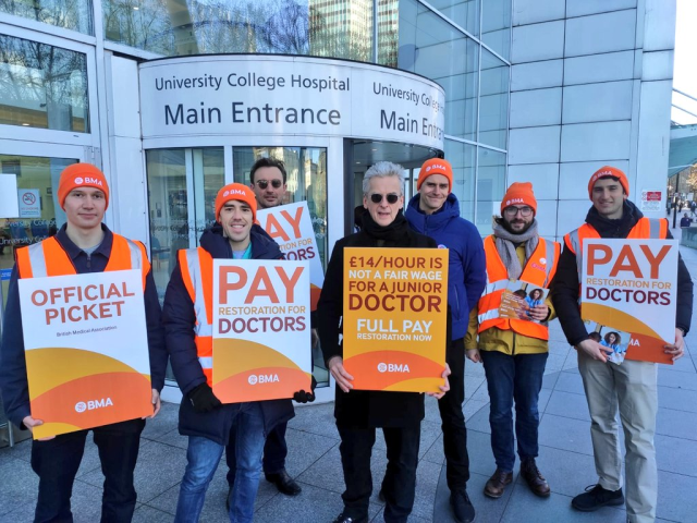 A picket line of several people wearing orange BMA clothes. Holding signs demanding pay restoration for doctors. Peter Capaldi, in black and sunglasses, stands at in the centre holding a sign saying "£14 / hour is not a fair wage for a junior doctor. Full pay restoration now"