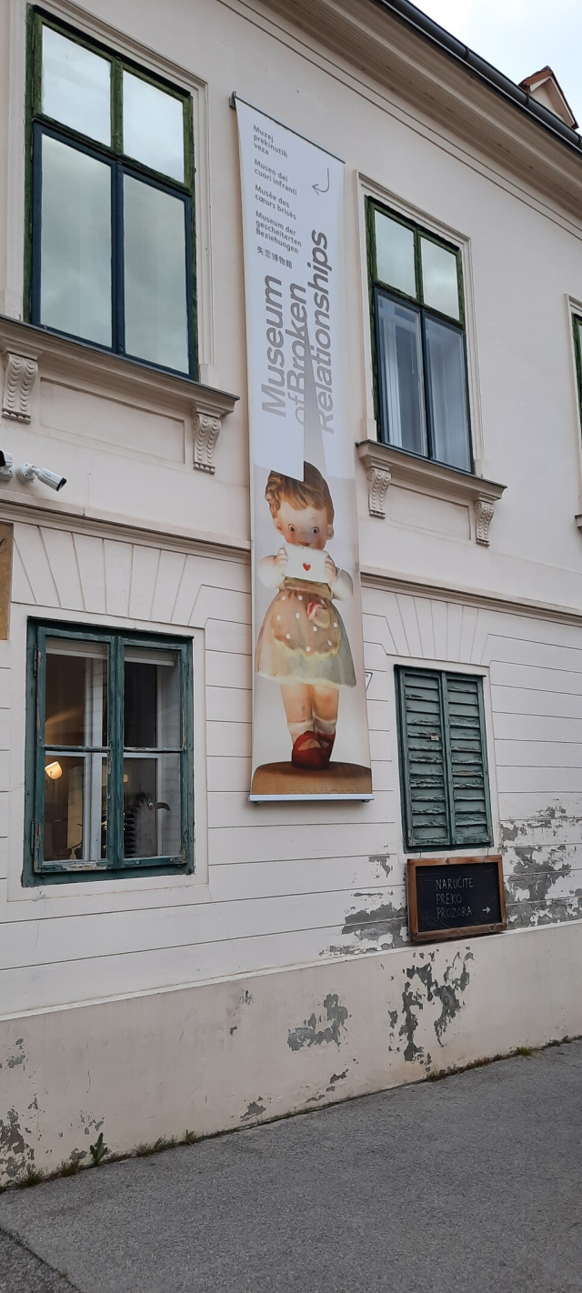 Photo of a banner on the side of a building, advertising the Museum of Broken Relationships. There is an image of a porcelain doll with a love letter in its hand.