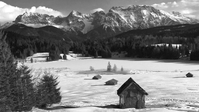 B&W photo of an alpine winter landscape, a view over Geroldsee towards the snow covered Karwendel massif with its steep rock faces. Several old hay barns/huts are standing in the alm meadows in the foreground. The lake surface is frozen and covered in snow too