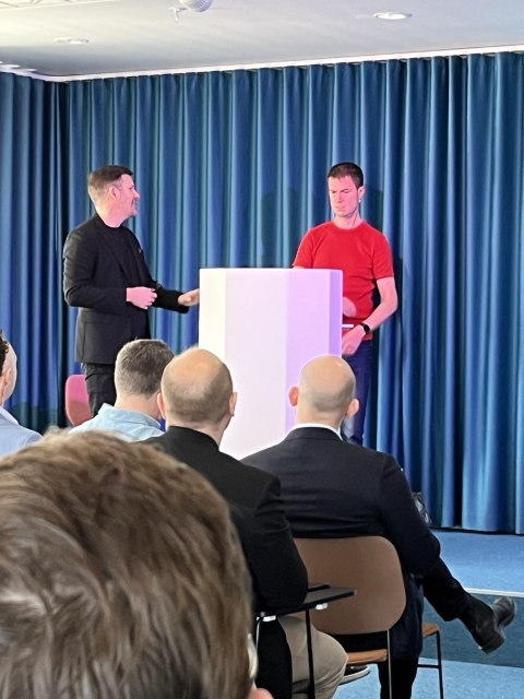 Carl Piva, CEO Internetstiftelsen (Swedish Internet Society) in a black suit introduces opening Keynote Speaker, Daniel Stenberg. Mr. Stenberg stands behind a white podium and the two gentlemen are in front of a dark blue curtain.