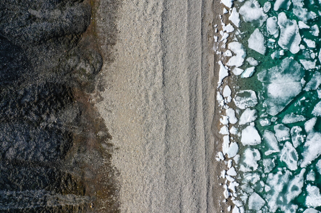 A drone shot looking down at a shoreline. Image is three almost equal parts. On the right is ice filled ocean, green/turquoise water. The middle is a gravelly beach. The left is tundra. 
