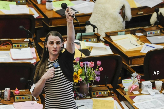 Rep. Zooey Zephyr holds her mic high in the air to protest her silencing by Montana House Republicans. Flowers offered by supporters cover the desk behind her. 