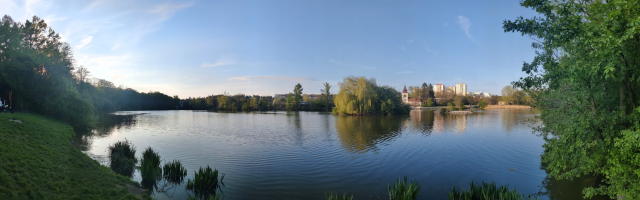 Panorama photo of a pond, with an church on the opposite bank and high density buildings in the background.