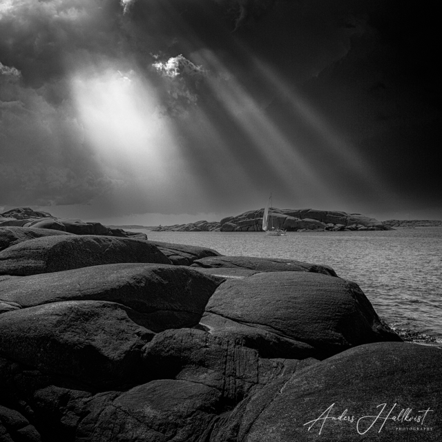 A black and white photo with rocks in the foreground then some sea water with a tiny sailing boat in front of a small island. The sky is very overcast and a few, strong light beams manage to get through the clouds.