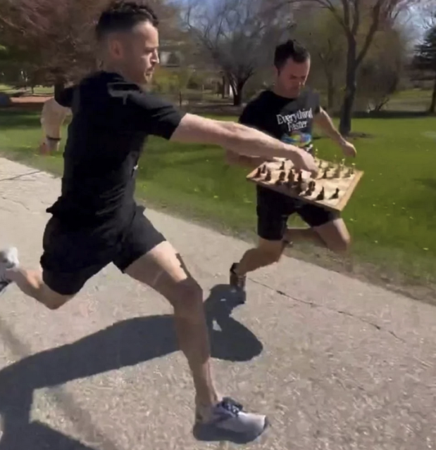 Two men running or jogging down a dirt road. One is holding up a chess board with pieces, and the other is making a move on the chess board.
