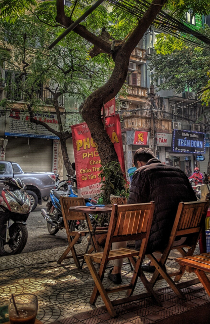 A man with his back to us sitting in a wooden chair is hunched over and looking at something in his hands or on his table. He's seated among several wooden chairs and tables arranged on a sun-dappled sidewalk—clearly the street-side seating of a small cafe. A tree rising from between the road and the tables provides shade. Motorscooters are parked along the curb, as is a silver pick-up on the opposite side of the street. Opposite the cafe is an unbroken line of narrow Hanoi rowhouses, each with first floor signs indicating the presence of various small, family businesses.