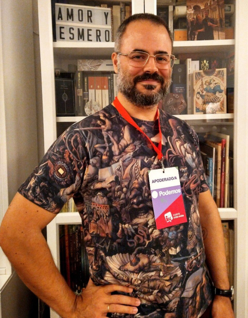 Foto de Yeray delante de una estantería llena de libros. Tiene una mano sobre la cadera, sonríe a cámara y en el cuello le cuelga una acreditación de apoderado por Podemos-Izquierda Unida. También lleva una camiseta super chula del Disco Elysium. Votad con responsabilidad el 28M y jugad al Disco Elysium también.