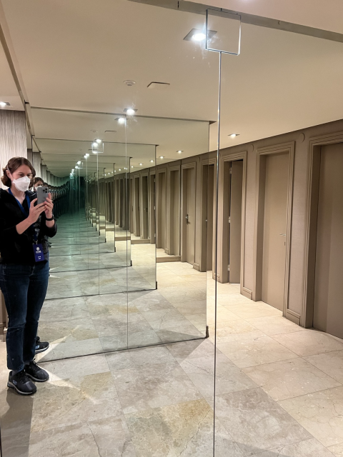 A shiny, marble-floored bathroom with mirrors set up so that it looks like the stalls extend off into the infinite distance. I'm standing to the side taking a photo with my phone, wearing a black sweater, jeans, and a white mask.