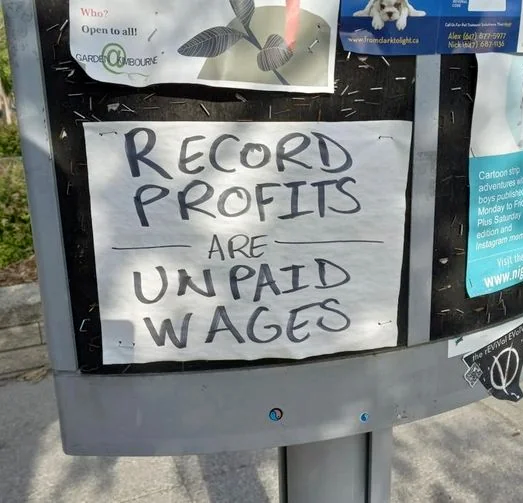 A handwritten sign posted to a bulletin board, reading "record profits are unpaid wages."