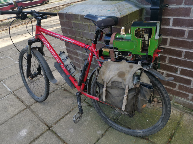A small green miniature steam locomotive strapped to the luggage rack of a push bike. The miniature railway can be seen in the background.