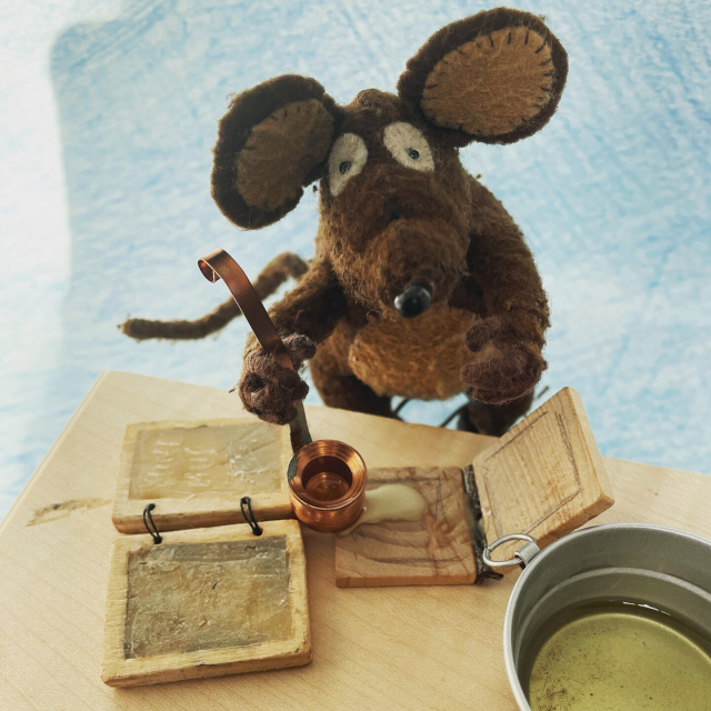 Photo of Minimus, the Latin mouse, pouring melted beeswax from a dipper onto a little hinged wooden writing tablet. There's a finished pair of tablets on the worktop next to him, and a pan of melted wax nearby