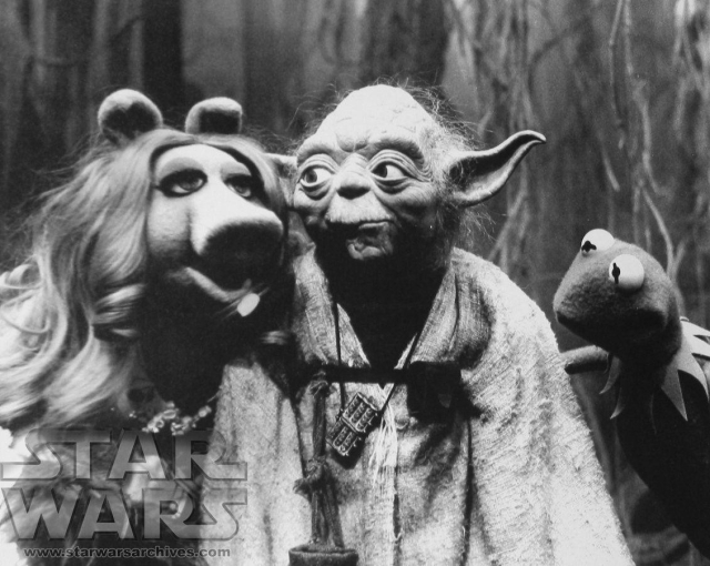 Black and white picture of Yoda and Miss Piggy, standing relatively close to each other, with Yoda turned to the left looking at Miss Piggy, and Kermit the frog on the other side looking at Yoda.