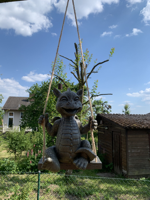 A small dragon statue sitting on a small swing visibly enjoying themselves. It's hanging from a metal support of an actual swing. There are some trees visible behind it.