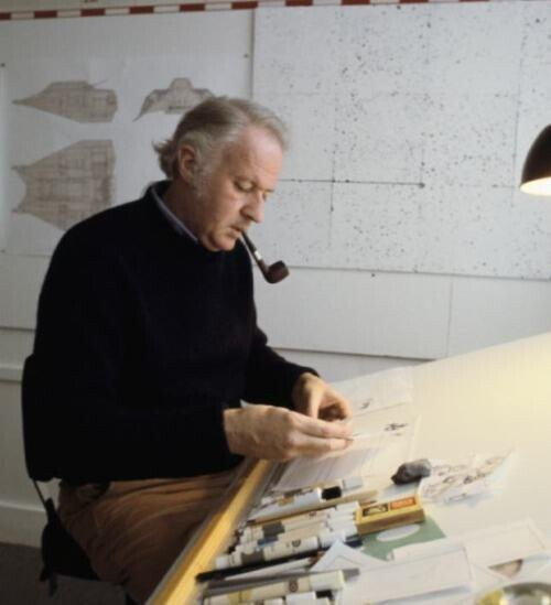 McQuarrie sitting at his drawing table. He is wearing a navy crew neck sweater and smoking a pipe. His hair is gray and swept back. There are numerous markers, pens, and erasers on the table, as well as a circle template and many scraps of paper.
