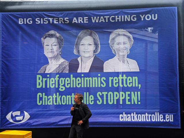 Ein Mann steht mit Mikro vor einem sehr großen Banner, auf dem Johansson, Feaser & von der Leye  steht: Big Sisters are Waschung you. Briefgeheimnis retten, Chatkontrolle stoppen! chatkontrolle.eu