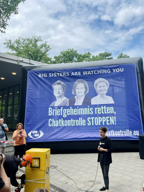 Foto einer Aktion gegen die Chatkontrolle. Im Hintergrund ein Banner mit den Gesichtern von EU Kommissarin Ylva Johannson, Bundesinnenministerin Nancy Faeser und EU Kommissionspräsidentin Ursula von der Leyen. 
Text: "Big sisters are watching you
Briefgeheimnis retten, 
Chatkontrolle STOPPEN!
chatkontrolle.eu" 
Im Vordergrund Piraten Vorstandsvorsitzende Anne Herperz mit Mikrofon. Neben ihr bricht ein Mann mit einem Werkzeug eine Kette auf, die um einen gelben Briefkasten gewickelt ist.