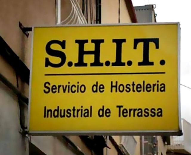 Black-on-yellow sign attached to a building for Servicio de Hosteleria Industrial de Terrassa (Terrassa Industrial Hospitality Service) with the acronym S.H.I.T. in much larger letters at the top
