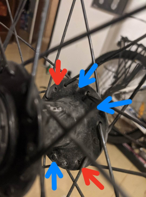 The detail photo of a rear bike wheel's free hub. There are 3 already replaced wires marked by blue arrow and distinguishable by wire head different from others, and there are 2 more wires marked by red arrows that are missing the head completely and needs replacement.